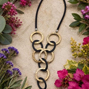 May include: A necklace featuring a black cord and a series of interlocking circular links. The design alternates between textured gold-toned metal rings and smooth matte black rings, arranged in a cascading vertical pattern that tapers downward.