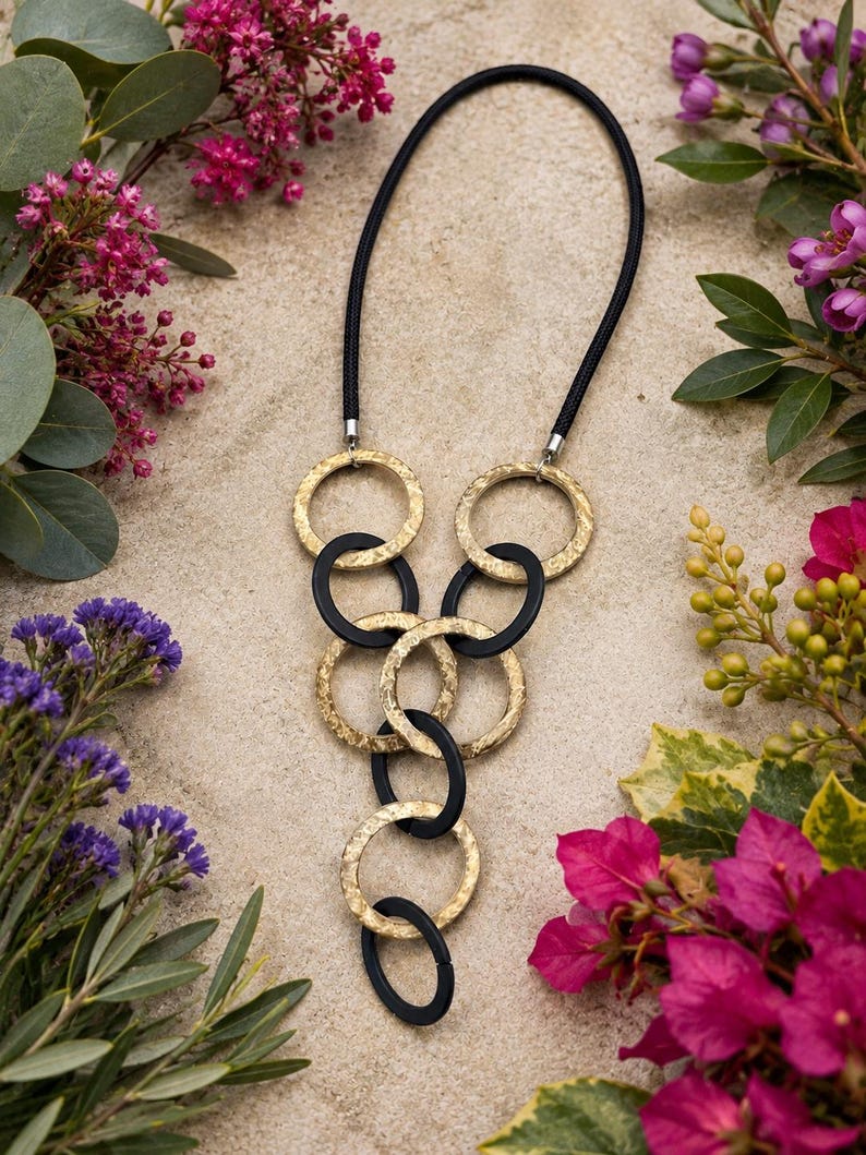 May include: A necklace featuring a black cord and a series of interlocking circular links. The design alternates between textured gold-toned metal rings and smooth matte black rings, arranged in a cascading vertical pattern that tapers downward.