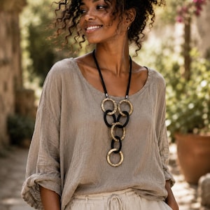 May include: A beige long-sleeved top with a scoop neckline and a necklace featuring alternating black and textured gold-toned circular rings linked together in a vertical drop design, suspended from a simple black cord.