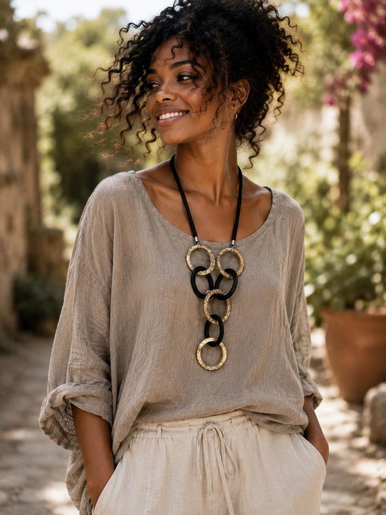 May include: A beige long-sleeved top with a scoop neckline and a necklace featuring alternating black and textured gold-toned circular rings linked together in a vertical drop design, suspended from a simple black cord.