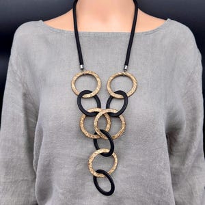 May include: A long necklace featuring a black cord and a series of interlocking circular pendants. The circles alternate between a textured, metallic gold finish and a smooth, matte black finish, arranged in a cascading, vertical pattern.