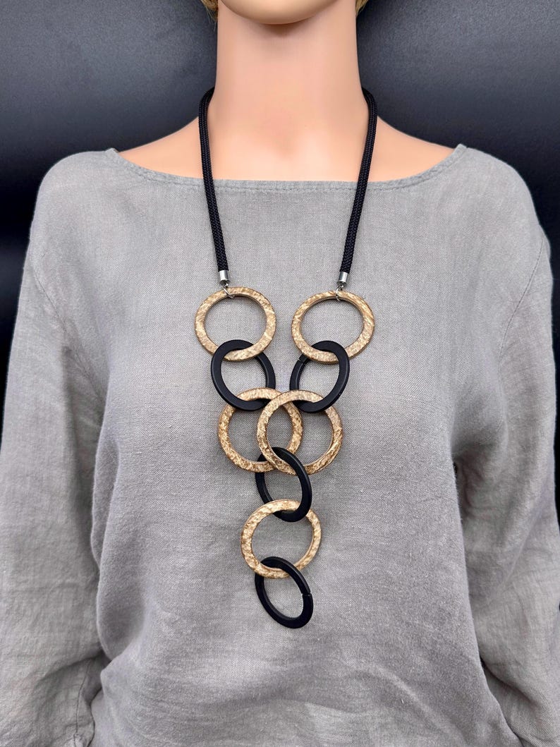 May include: A long necklace featuring a black cord and a series of interlocking circular pendants. The circles alternate between a textured, metallic gold finish and a smooth, matte black finish, arranged in a cascading, vertical pattern.