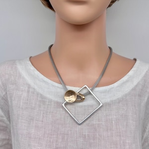 May include: A silver and gold necklace with a geometric pendant. The pendant is a square with rounded corners and a small circle attached to it. The necklace is on a grey cord.