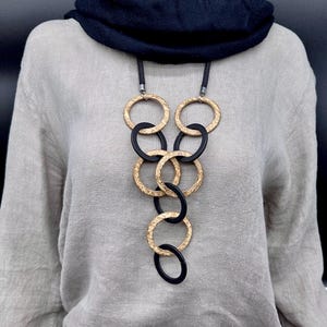 May include: A long necklace featuring a series of interlocking circular rings in alternating gold-tone textured metal and smooth matte black material, suspended from a thick black cord, worn over a light gray long-sleeve top and a black scarf.