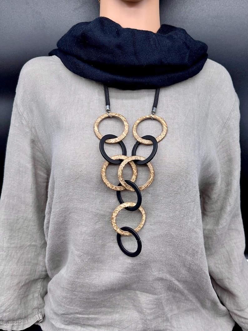 May include: A long necklace featuring a series of interlocking circular rings in alternating gold-tone textured metal and smooth matte black material, suspended from a thick black cord, worn over a light gray long-sleeve top and a black scarf.