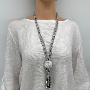 Könnte beinhalten: Eine lange silberne Perlenkette mit einem großen weißen Kugelanhänger und einem silbernen Quastenanhänger.