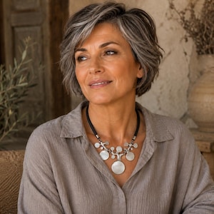 Peut inclure: Un collier imposant composé d'un cordon noir et de plusieurs disques métalliques argentés de tailles variées disposés en forme de branches. Le collier repose sur une chemise à col gris clair portée par une personne aux cheveux courts gris.