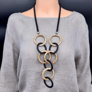 May include: A long necklace featuring a black cord and a series of interlocking circular rings. The rings alternate between a textured, metallic gold finish and a smooth, matte black finish, arranged in a cascading vertical pattern.