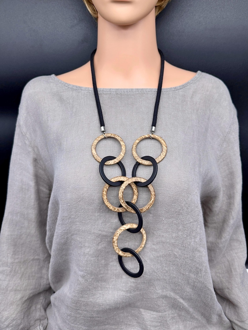 May include: A long necklace featuring a black cord and a series of interlocking circular rings. The rings alternate between a textured, metallic gold finish and a smooth, matte black finish, arranged in a cascading vertical pattern.
