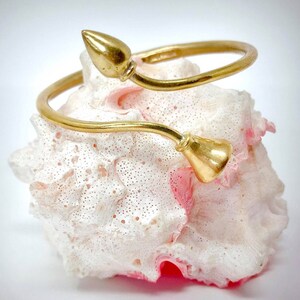 Op de afbeelding: Een goudkleurige, open armband met een bloemmotief. De armband heeft aan elk uiteinde een knopvormig element en wordt gepresenteerd op een wit en roze koraalachtig oppervlak. Het ontwerp van de armband is eenvoudig en elegant.
