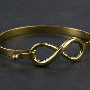 Op de afbeelding: Een goudkleurige armband met een oneindigheidssymbool. De armband heeft een sluiting en is gemaakt van een glanzend, metalen materiaal. Het oneindigheidssymbool is een prominent ontwerpelement.