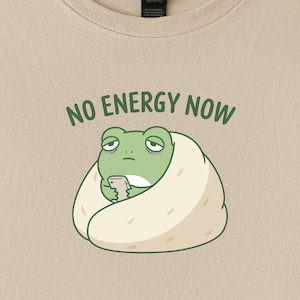 May include: Beige t-shirt featuring a cartoon frog wrapped in a burrito, holding a phone. The text "NO ENERGY NOW" is above the frog in green letters. The frog has a tired expression.