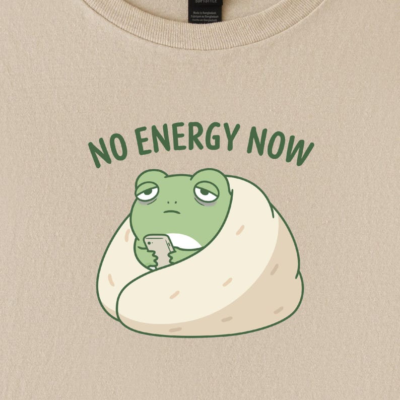 May include: Beige t-shirt featuring a cartoon frog wrapped in a burrito, holding a phone. The text "NO ENERGY NOW" is above the frog in green letters. The frog has a tired expression.