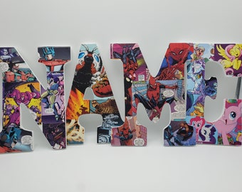 May include: White wooden letters spelling out the word "NAME" decorated with colorful comic book character images. The letters are decorated with images of Spiderman, Transformers, My Little Pony, and other comic book characters.