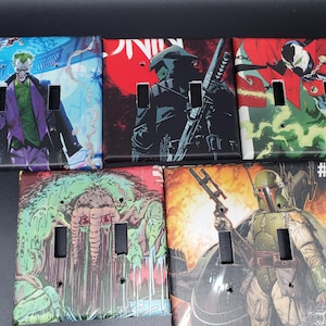 May include: Five light switch covers with comic book art. The covers feature characters like the Joker, a ninja, Spawn, a monster, and Boba Fett. Each cover has two rectangular cutouts for the switches and screw holes.