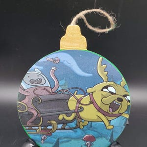 May include: A round, flat, green-edged ornament featuring a cartoon illustration of a yellow dog with antlers pulling a dark sleigh with a pink character inside. The scene is set against a blue winter landscape with trees. A gold cap with a twine loop is attached.