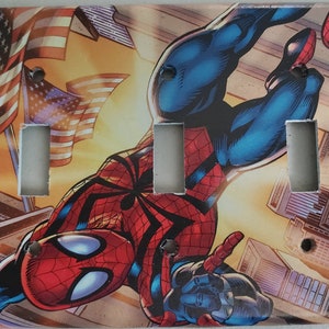 May include: A decorative light switch cover featuring a comic book illustration of Spider-Man in a dynamic pose. The background includes American flags and city buildings. The cover has three switch openings and screw holes.
