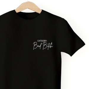 May include: Black t-shirt with the text "CATEGORY: Bad Bitch" in white cursive lettering.