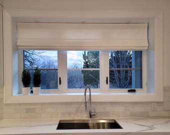 May include: A white kitchen with a stainless steel sink and a window with white roman shades. The window has a view of trees and a cloudy sky.