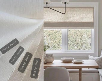 May include: A white linen Roman shade with a black metal rod, hanging in a window with a view of trees. The shade is raised, revealing the window behind it. The window is framed in white trim. The shade is available in five colors: Comfy White, Comfy Cream, Comfy Bone, Comfy Fog, and Comfy Linen.