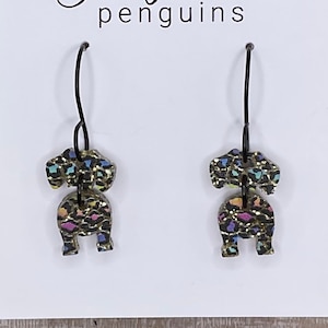 May include: A pair of dangle earrings featuring a black and gold leopard print design with pops of pink, blue, and yellow. The earrings are shaped like a penguin and hang from black hooks. The earrings are displayed on a white card with the text "eighty-four penguins".