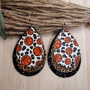 May include: A pair of dangle earrings with a layered design. The top layer features a white background with orange basketballs and black leopard print. The middle layer is a brown leopard print. The bottom layer is black glitter.