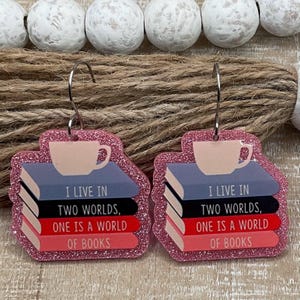 May include: Pink glittery earrings with a book and coffee cup design. Each earring features a stack of books in blue, black, and red, topped with a light pink coffee cup. The text reads "I LIVE IN TWO WORLDS, ONE IS A WORLD OF BOOKS."