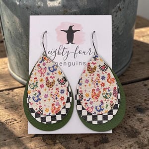 May include: Teardrop-shaped layered earrings featuring a white top layer with a colorful chicken and flower pattern, a middle layer with a black and white checkered border, and a solid olive green bottom layer with silver-toned metal hooks.