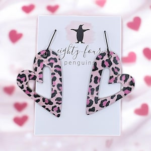 May include: Pink and black leopard print heart-shaped earrings with a cutout heart design. The earrings are on a white card with the text "eighty-four penguins".