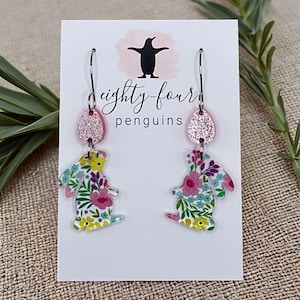 May include: A pair of dangle earrings featuring a bunny design with a floral pattern in pink, yellow, and green. The earrings have a pink glitter accent and are attached to a silver-tone hook. The earrings are displayed on a white card with the text "eighty-four penguins".