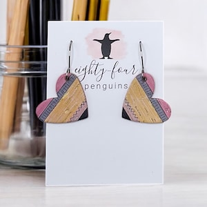 May include: Heart-shaped earrings with a pink, yellow, and black design. The earrings are on a white card with the text "eighty-four penguins" and a penguin logo. The earrings have a unique, textured appearance.