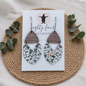 May include: A pair of dangle earrings with a wooden triangle top and a heart-shaped bottom. The heart-shaped bottom is white with black school-themed illustrations. The earrings hang from silver-colored hooks. The earrings are displayed on a white card with the text "eighty-four penguins".