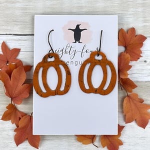 May include: A pair of orange pumpkin-shaped earrings with black hooks. The earrings are made of a textured material and are displayed on a white card with the text "Eighty-four Penguins". Autumn leaves surround the card.