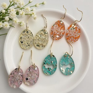 May include: Six pairs of egg-shaped earrings in various colors and designs. The earrings have gold-colored hooks. Colors include gold, orange, pink, and teal. Some earrings have glitter accents, and some have cut-out designs.