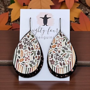 May include: Teardrop-shaped earrings with a layered design. The top layer features a cream background with a botanical print of leaves and berries. The middle layer has vertical stripes, and the bottom layer is a dark brown glitter. The earrings are displayed on a white card with the text "eighty-four penguins".