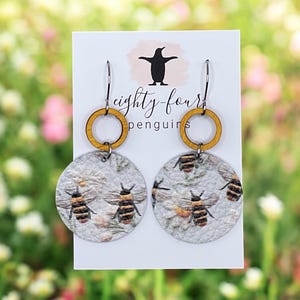 May include: A pair of round earrings featuring a bee design. The earrings have a light gray background with a floral pattern and several bees. The top of the earrings have a wooden ring and a silver hook. The earrings are displayed on a white card with the text "eighty-four penguins".