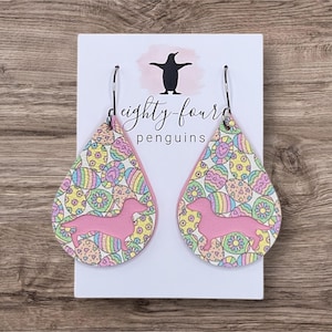 May include: A pair of pink teardrop-shaped earrings with a white background and a pastel Easter egg pattern. The earrings feature a pink dachshund silhouette.