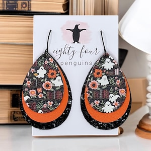 May include: Teardrop-shaped earrings with a Halloween theme. The earrings feature a black glitter base, an orange middle layer, and a top layer with a floral and ghost pattern. The text "eighty-four penguins" is visible on the backing card.