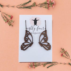 May include: Wooden butterfly wing earrings displayed on a white card with the text "eighty-four penguins". The earrings are brown with intricate cut-out details. The background is a soft peach color, with sprigs of greenery and pink flowers.