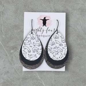 May include: A pair of dangle earrings with a black and white dachshund pattern. The earrings are made of leather and have a silver glitter finish. The earrings are on a white card with the text "eighty-four penguins".