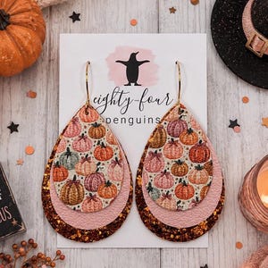 May include: Teardrop-shaped earrings featuring a layered design with a top layer of pumpkin-patterned material, a middle layer of light pink textured material, and a bottom layer of dark copper glitter, suspended from gold-toned metal hooks.
