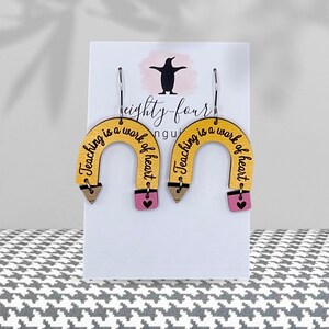 May include: A pair of wooden earrings shaped like pencils. The earrings are yellow with a brown pencil tip, a pink eraser, and the words "Teaching is a work of heart" in black. The earrings hang from silver hooks.