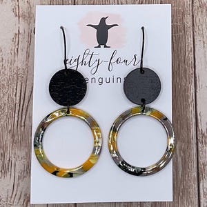 May include: A pair of dangle earrings featuring a black wooden disc above a large, circular hoop. The hoops have a marbled pattern in shades of yellow, gray, and black. The earrings are displayed on a white card with a penguin logo and the text "eighty-four penguins".
