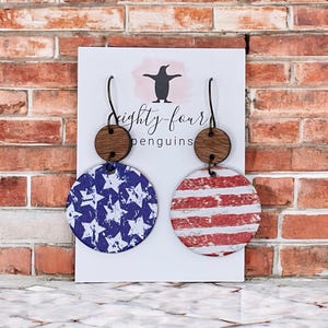 May include: A pair of patriotic earrings featuring a wooden top and a circular design. One earring displays a blue background with white stars, while the other showcases red and white stripes. The earrings are displayed on a white card with the text "eighty-four penguins".