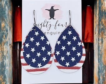 May include: Patriotic teardrop earrings featuring a layered design. The top layer displays a navy blue background with white stars, while the bottom layer showcases red and white stripes. The earrings are displayed on a white card with the text "eighty-four penguins".