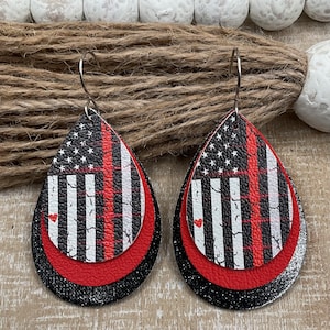 May include: A pair of dangle earrings with a layered design. The top layer features a distressed American flag with a red stripe down the middle. The middle layer is a solid red circle. The bottom layer is a black circle with silver glitter.