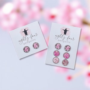 May include: Two sets of stud earrings on white cards. One set features pink ribbon designs on a leopard print background. The other set includes pink glitter and solid pink studs. The cards have the text "eighty-four penguins" and a penguin logo.