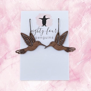 May include: Wooden hummingbird earrings with intricate wing details. The earrings are a warm brown color and hang from thin, dark brown wires. The earrings are displayed on a white card with the text "eighty four penguins" and a penguin logo, set against a pink background.
