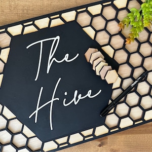 以下が含まれることがあります： ハニカム模様の黒い木製看板に、「The Hive」という文字が白く書かれています。看板は木製の表面に置かれ、背景には小さな多肉植物があります。