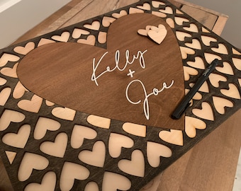Könnte beinhalten: Ein Gästebuch aus Holz in Herzform mit einem großen Herz-Ausschnitt in der Mitte. Das ausgeschnittene Herz ist braun und trägt die Namen "Kelly + Joe" in weißer Schrift. Der umgebende Bereich ist mit kleineren Herz-Ausschnitten bedeckt, auf denen ein schwarzer Stift liegt.